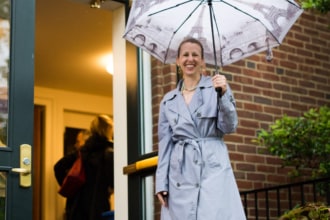 A woman in a coat holding an umbrella stands in front of open doors.