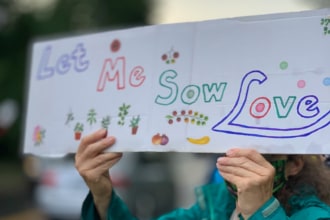 Hands holds a hand drawn protest sign with the images of plants and the words: let me sow love.